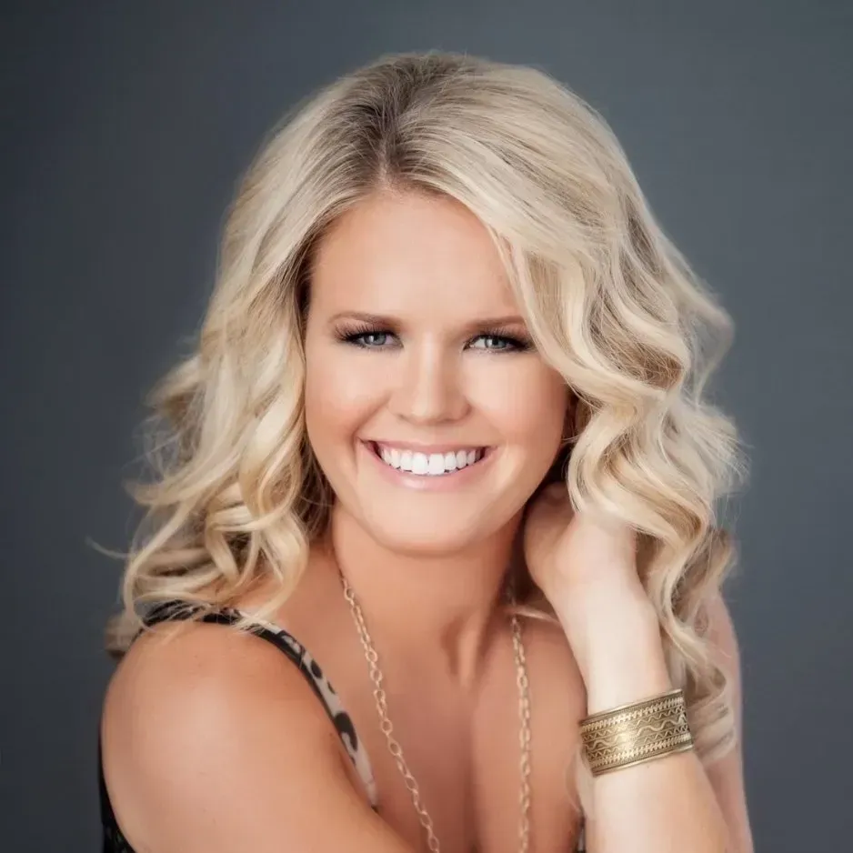 Blonde woman smiles, resting hand near wavy hair, wearing gold jewelry, against a gray background.
