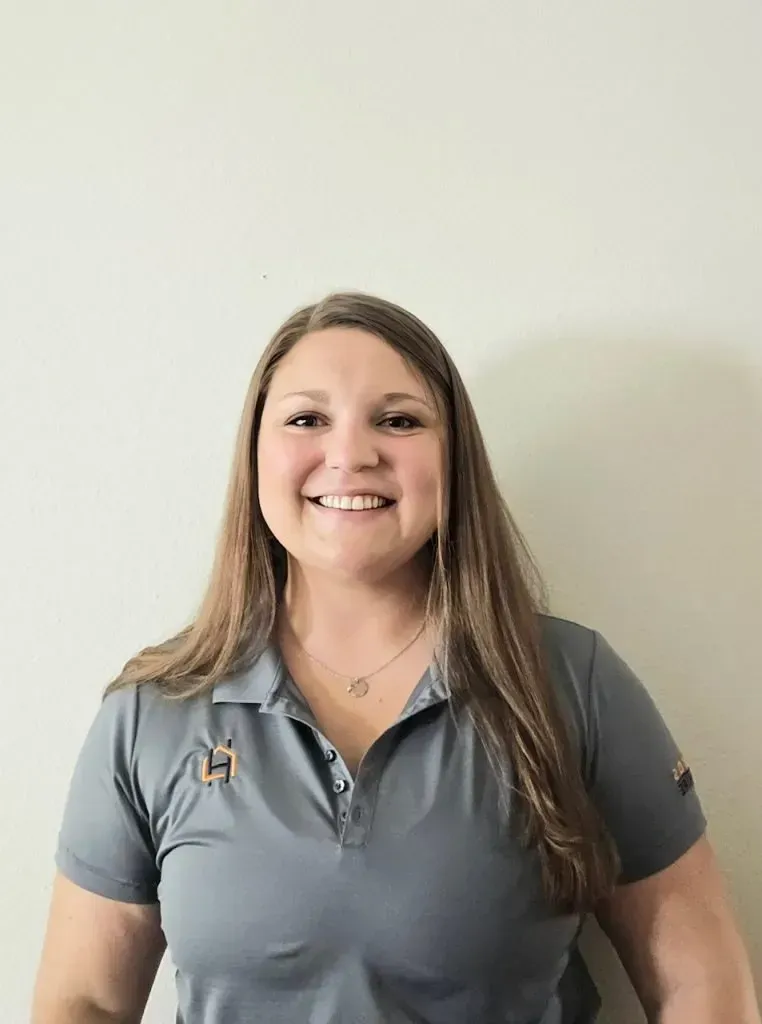 Woman with long brown hair wearing a gray polo shirt, smiling in front of a neutral wall.