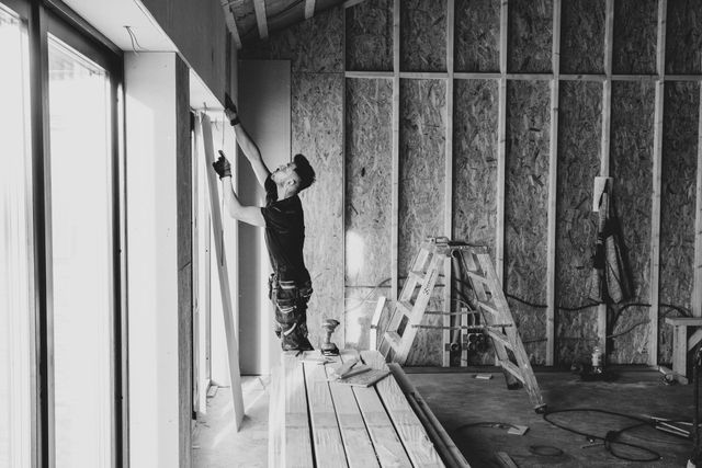 Person installing drywall near a large window in a wood-framed building under construction.