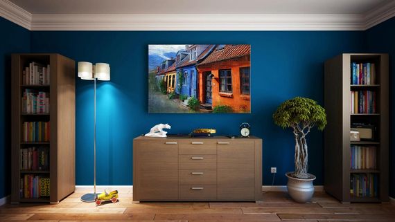 A room with a blue wall, painting, bookshelves, and a wooden cabinet with a lamp and a plant.
