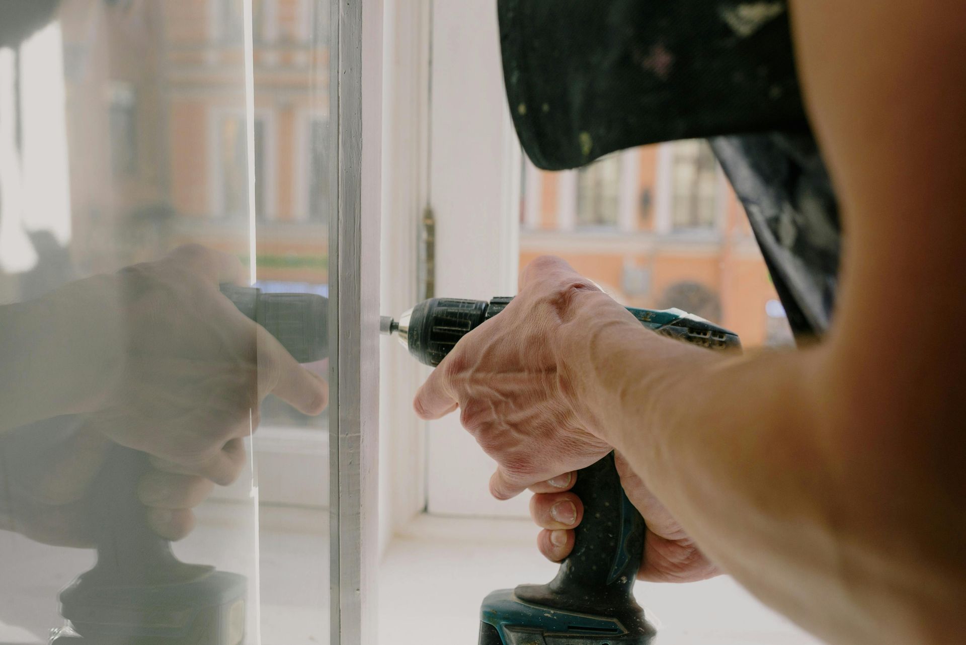 Person using a power drill on a white window frame near a glass pane, working indoors.