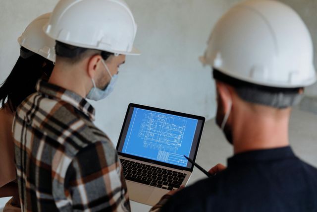 Construction workers in hard hats and masks review blueprints on a laptop.