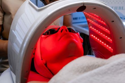 Person under a red light therapy panel wearing protective goggles and a towel-around neck.