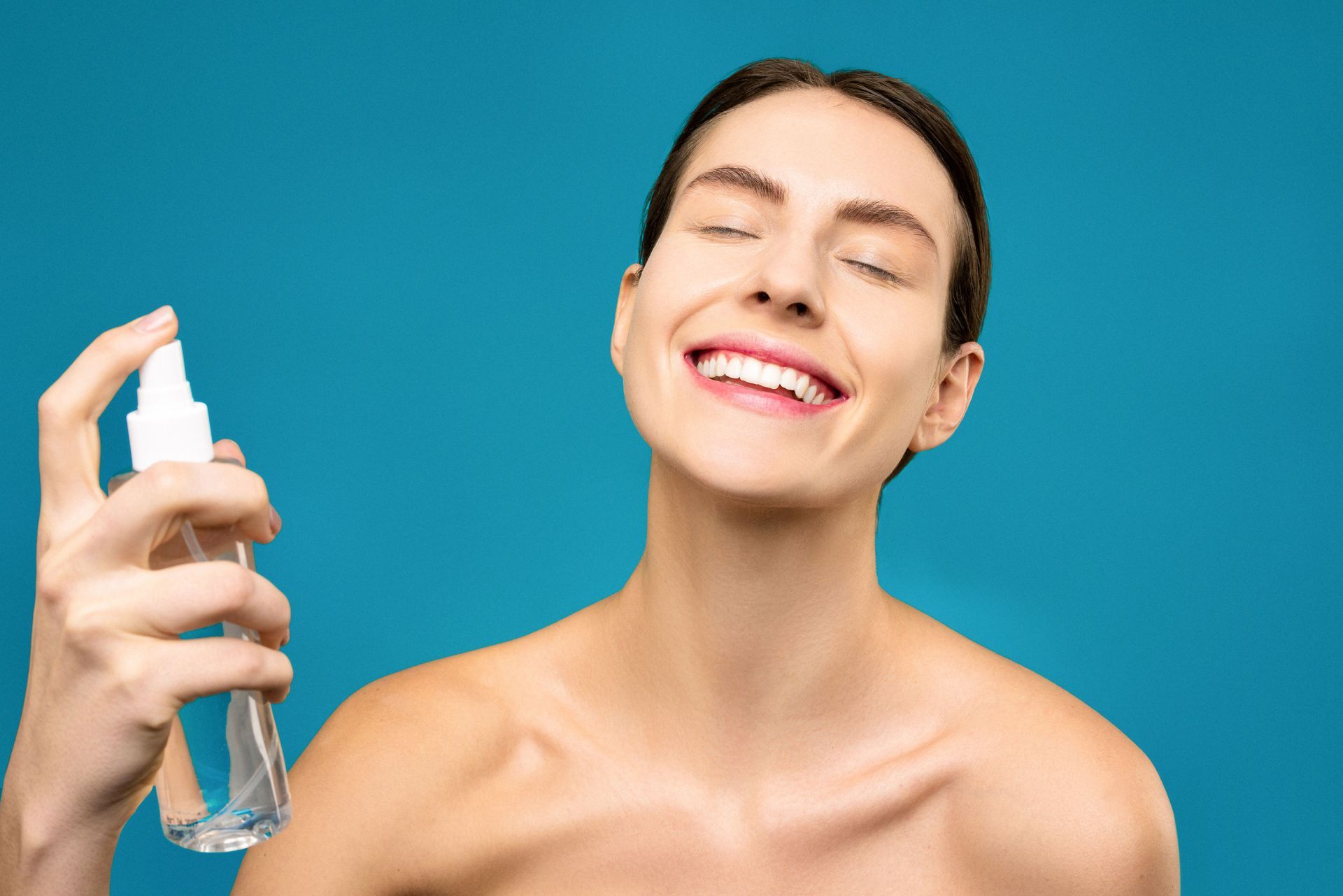Smiling person spraying mist on their face against a blue background