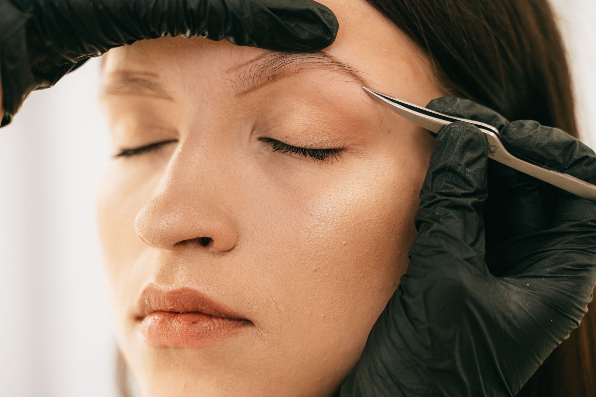 Gloved hands applying eyebrow makeup to a woman with closed eyes