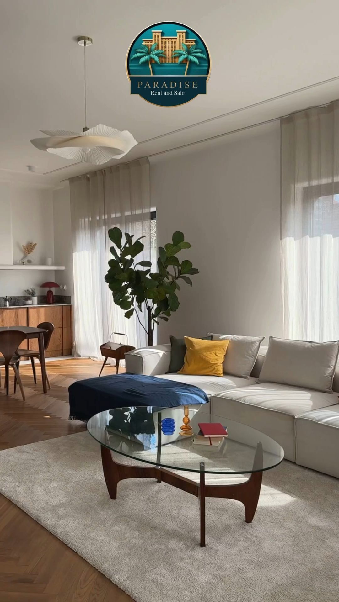 Sala de estar moderna con sofá blanco, mesa de centro de cristal, otomana azul, plantas y cortinas transparentes.
