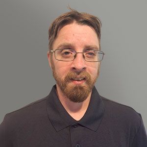 Portrait of a man with glasses and a beard wearing a dark collared shirt against a gray background