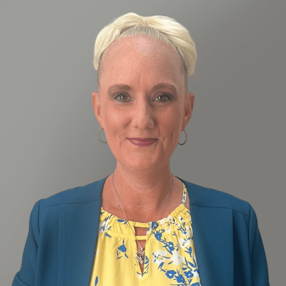Smiling woman in a blue blazer and yellow floral top against a gray background