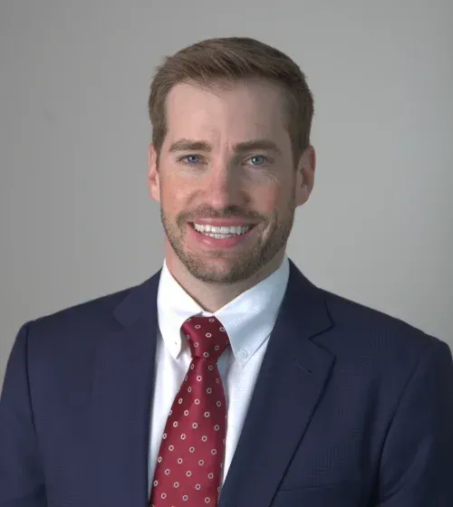 Portrait of a smiling man in a navy suit, white shirt, and red patterned tie against a gray background