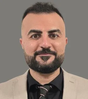 Portrait of a man with a beard in a beige blazer, black shirt, and striped tie against a gray background