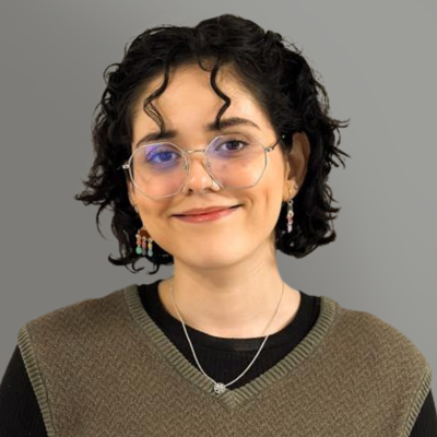 Portrait of a smiling person with short curly hair, glasses, and a brown sweater against a gray background