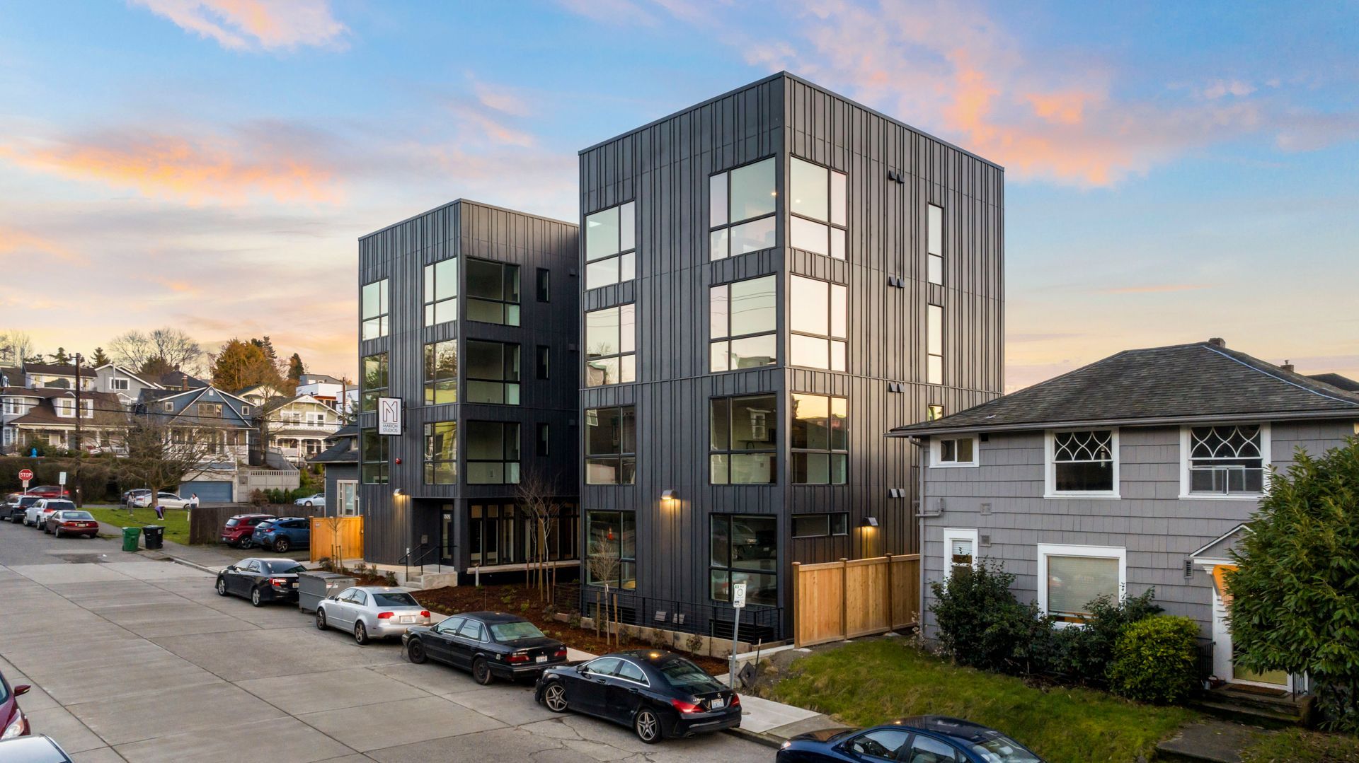 Modern black apartment building on a sloped street with parked cars at sunset
