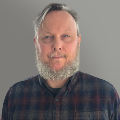 Portrait of a bearded man in a dark plaid shirt against a plain gray background