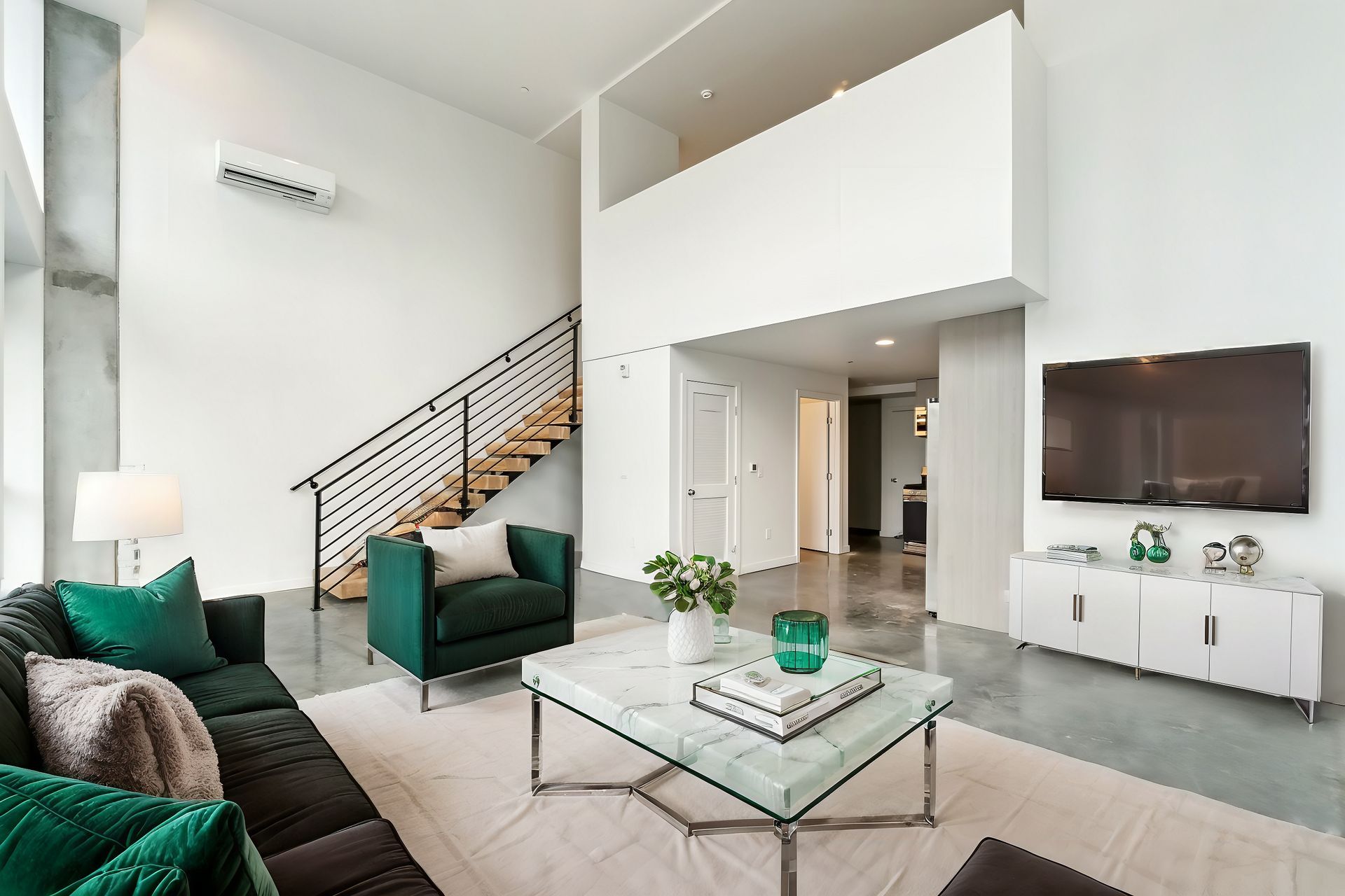 Bright modern living room with white walls, green chairs, glass coffee table, staircase, and TV console