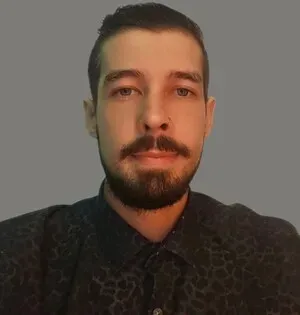 Portrait of a man with short dark hair and beard, wearing a dark patterned shirt against a gray background