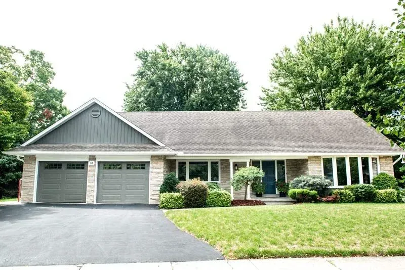 Ranch-style house with gray garage doors, brick facade, and a blue front door, set on a green lawn with trees in the background.