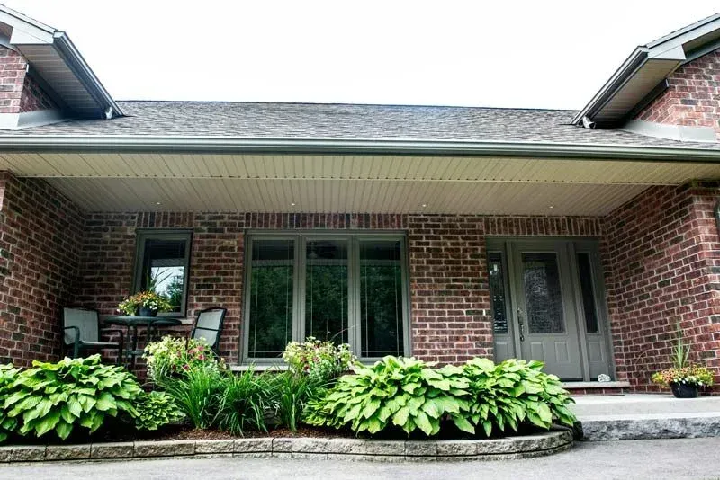 Red brick house with a covered porch. Large windows and a front door, with green plants in front.