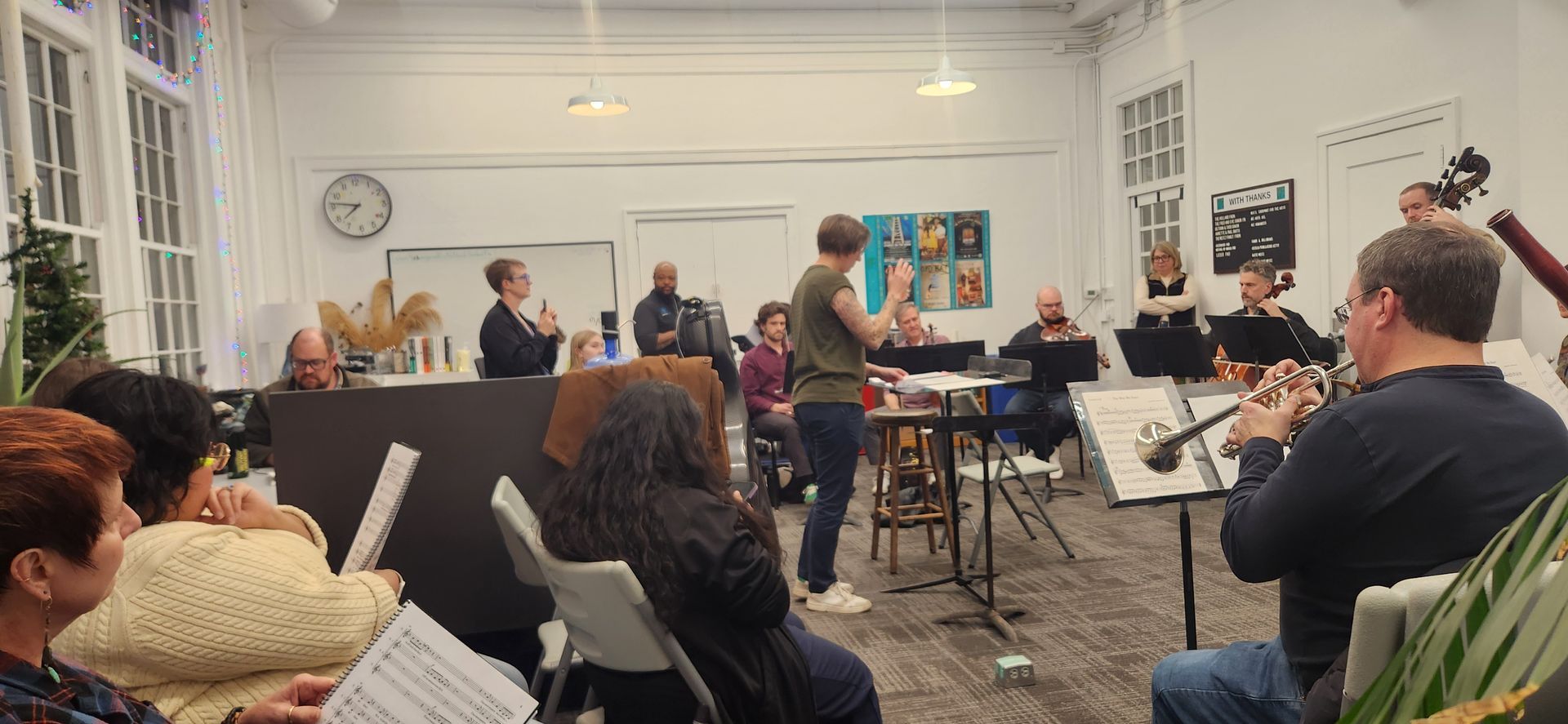 Musicians rehearsing in a bright room with seated audience members and music stands.