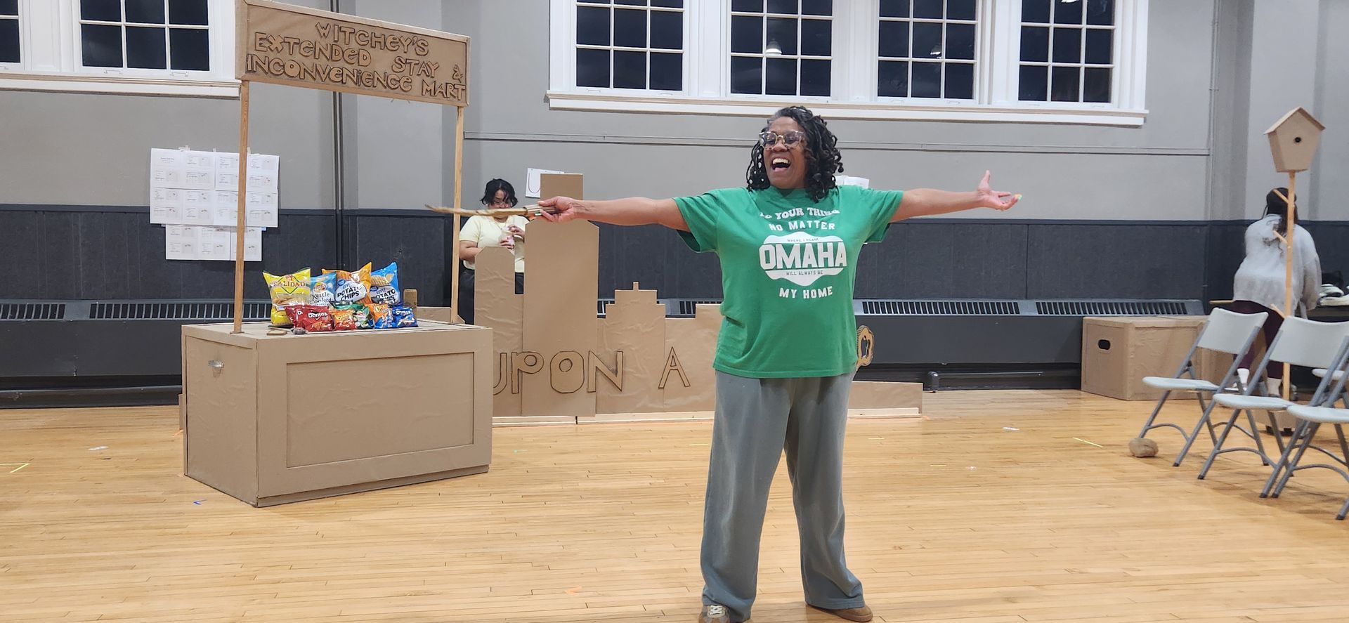 Person in a green T-shirt stands with arms outstretched in a gym-like room with tables and boxes.