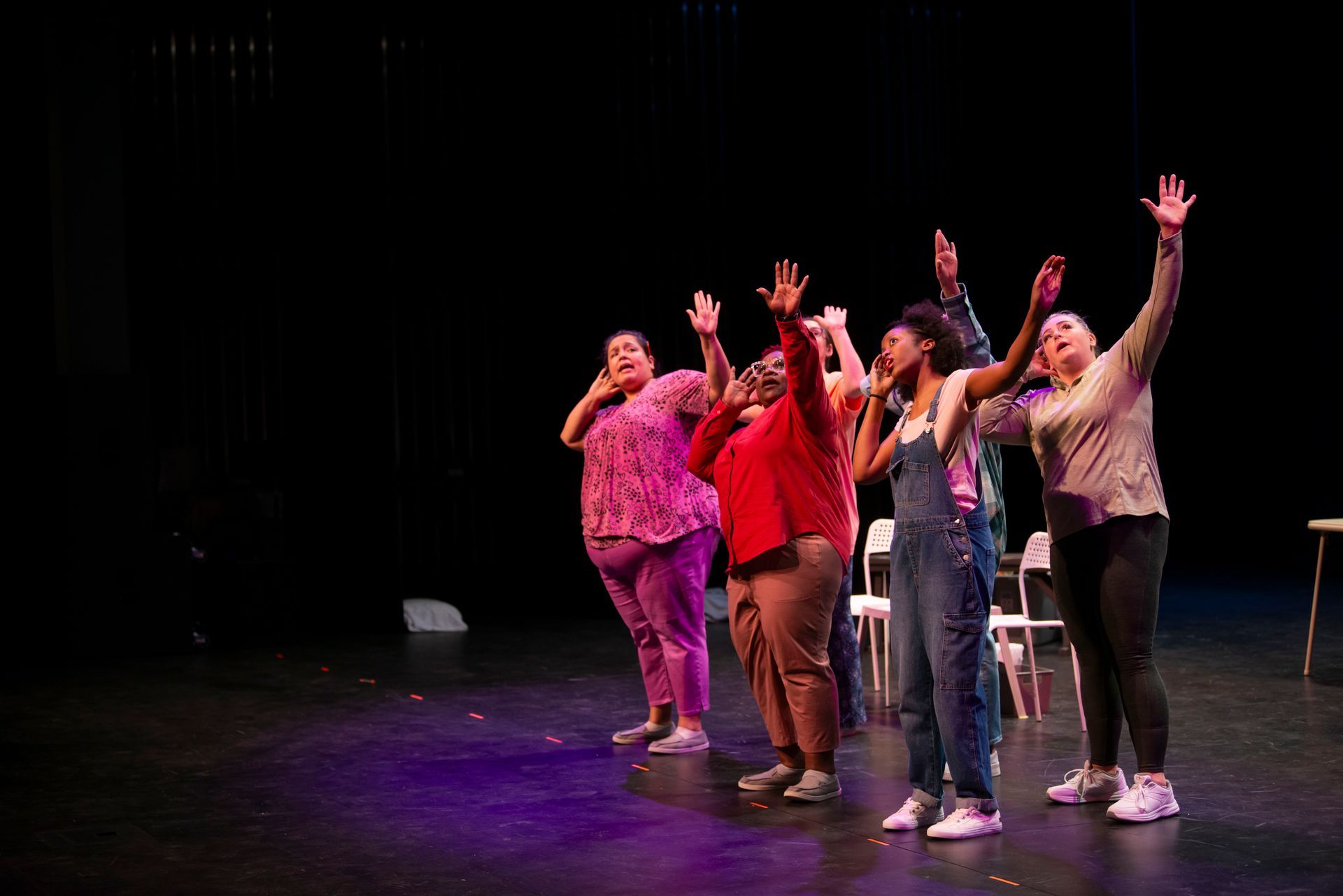 Five performers on a dark stage, posing with arms raised and colorful outfits under stage lights