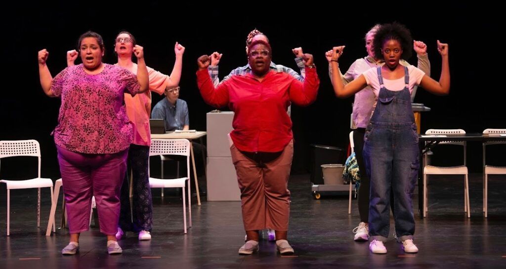 Three performers on stage, arms raised, in colorful outfits with chairs in the background