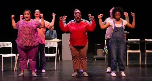 Three performers on stage, arms raised, in colorful outfits with chairs in the background