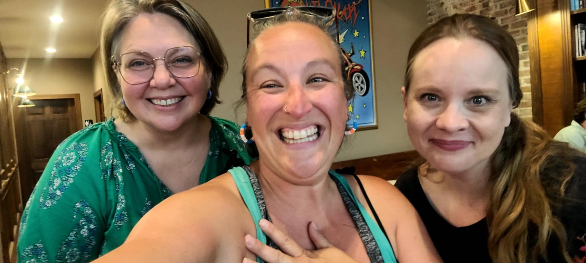 Three smiling women pose together indoors, with warm wood-paneled background.