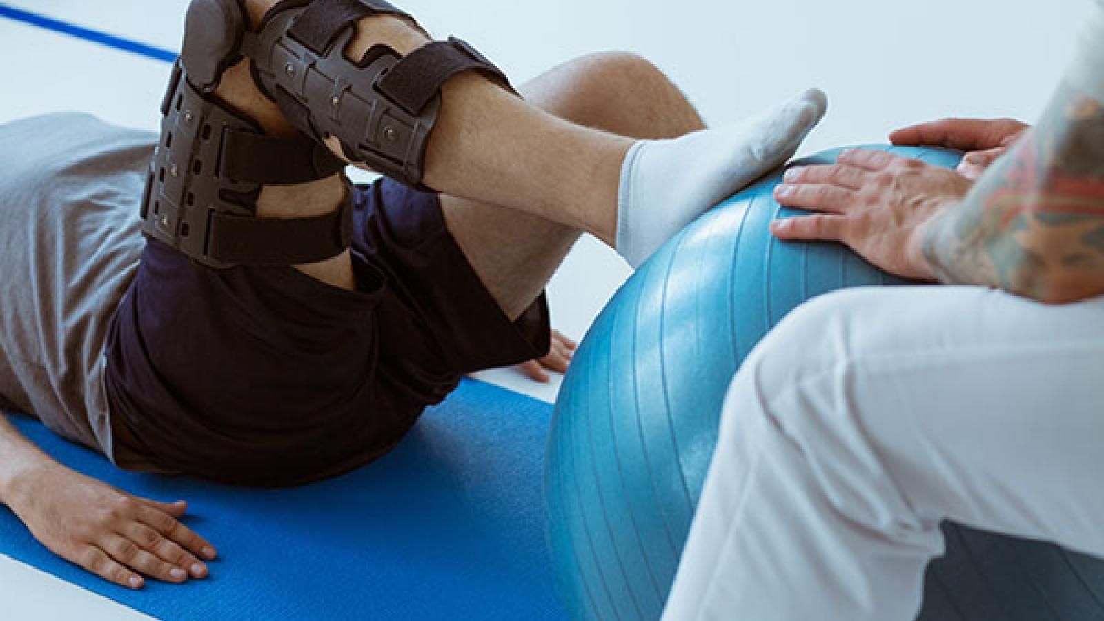A person with a knee brace is laying on a yoga mat next to an exercise ball.