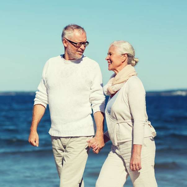 Couple holding hands, smiling at each other while walking on a beach. Ocean and blue sky background.