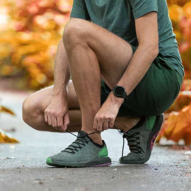 Person in green athletic wear tying running shoe on a paved path, watch on wrist, autumn foliage.