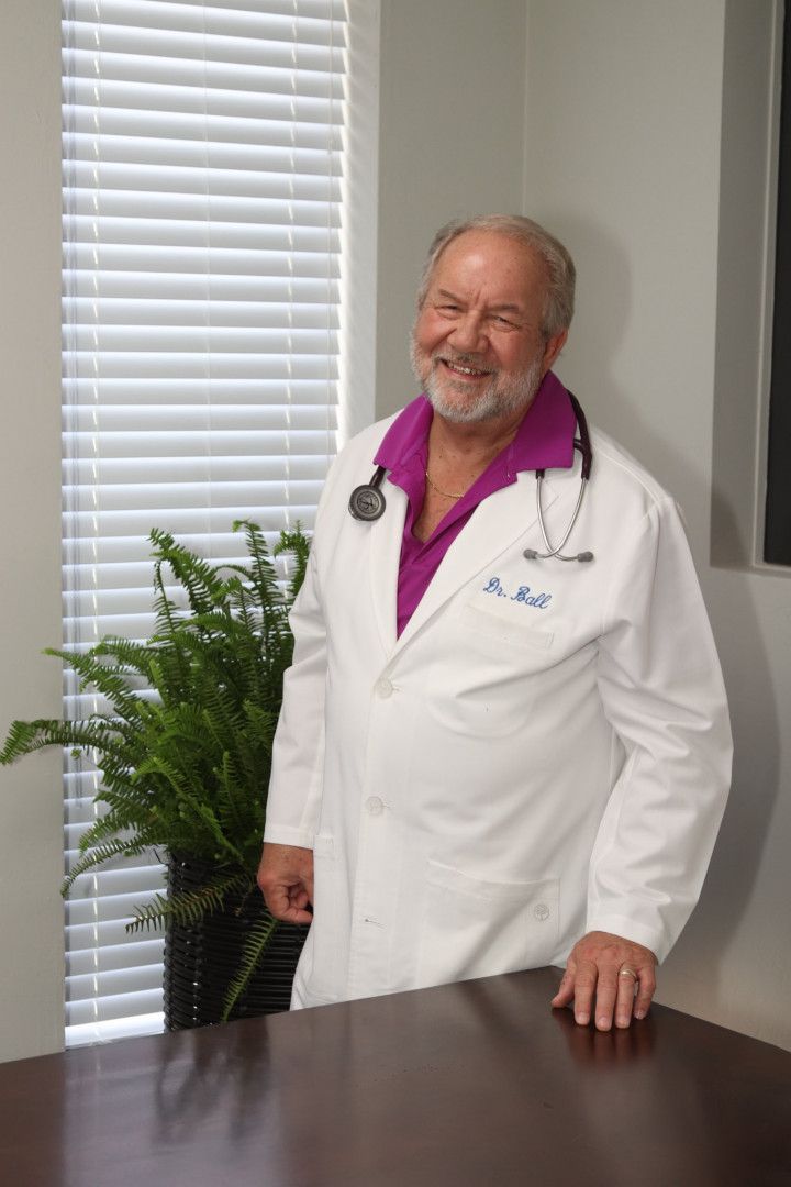 Doctor with stethoscope smiles at camera, wearing a white coat over a purple shirt, office setting.