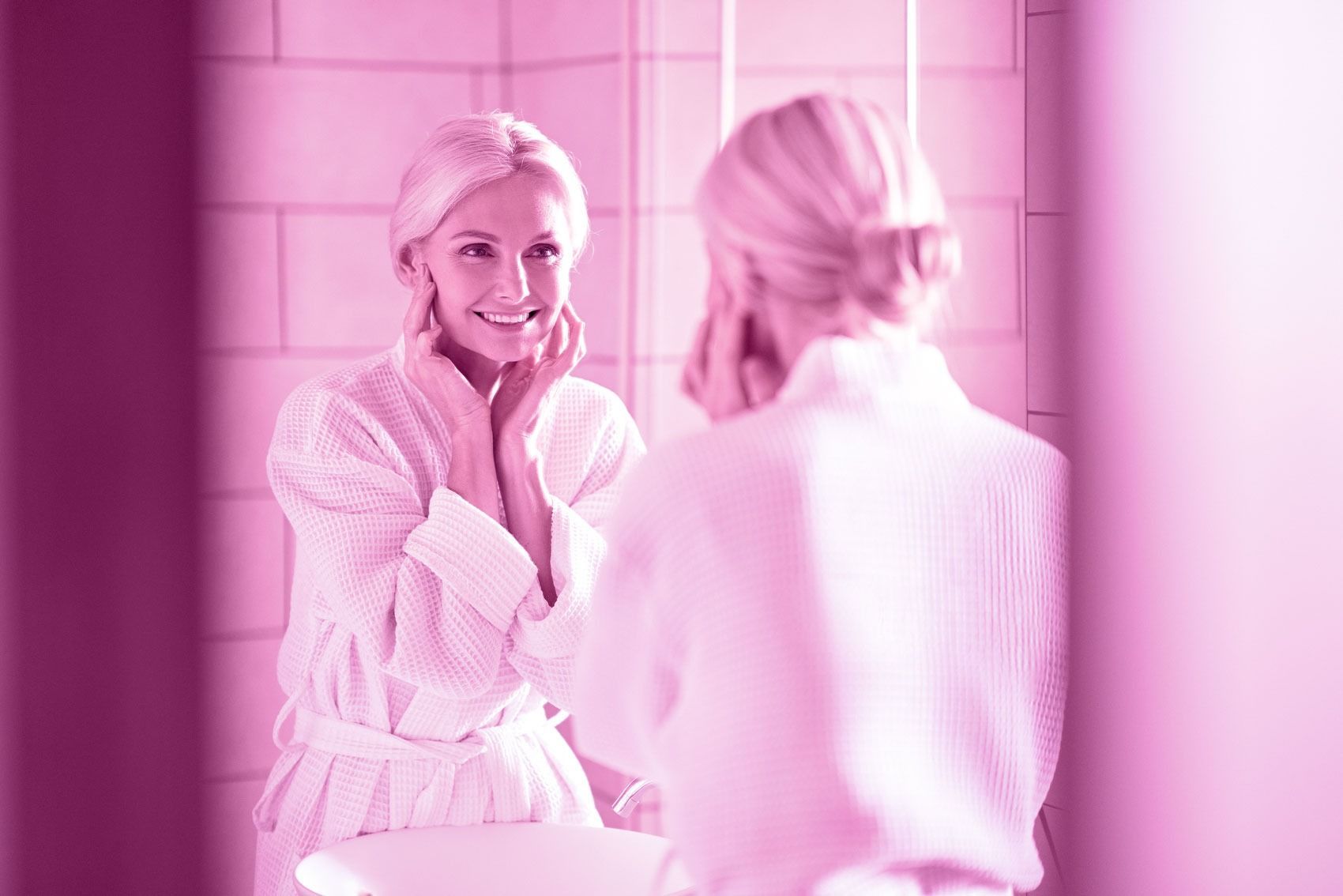 Woman in a robe looking in the mirror, touching her face, smiling in a pink-toned bathroom.