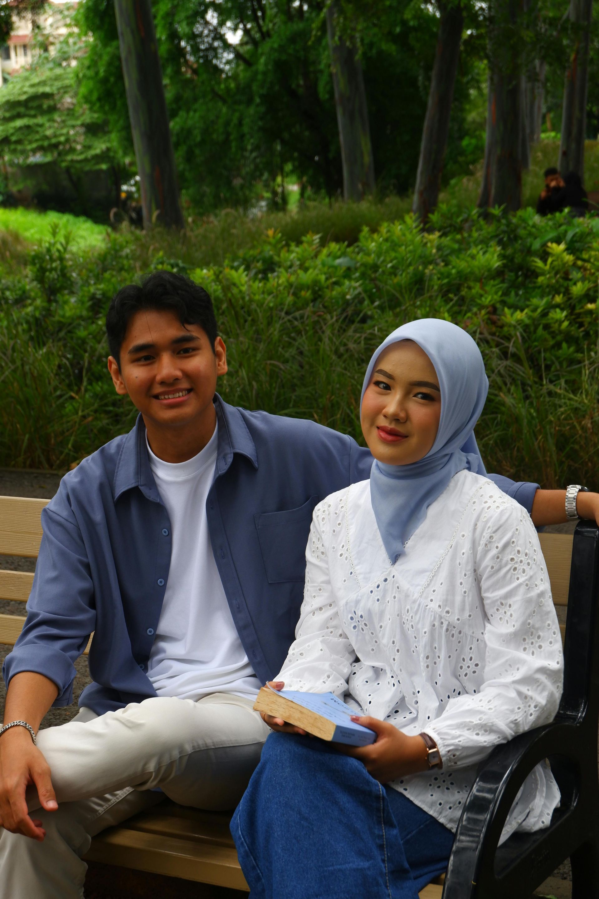 Couple seated on a park bench. Man in blue shirt, woman in hijab and white shirt, holding a book.