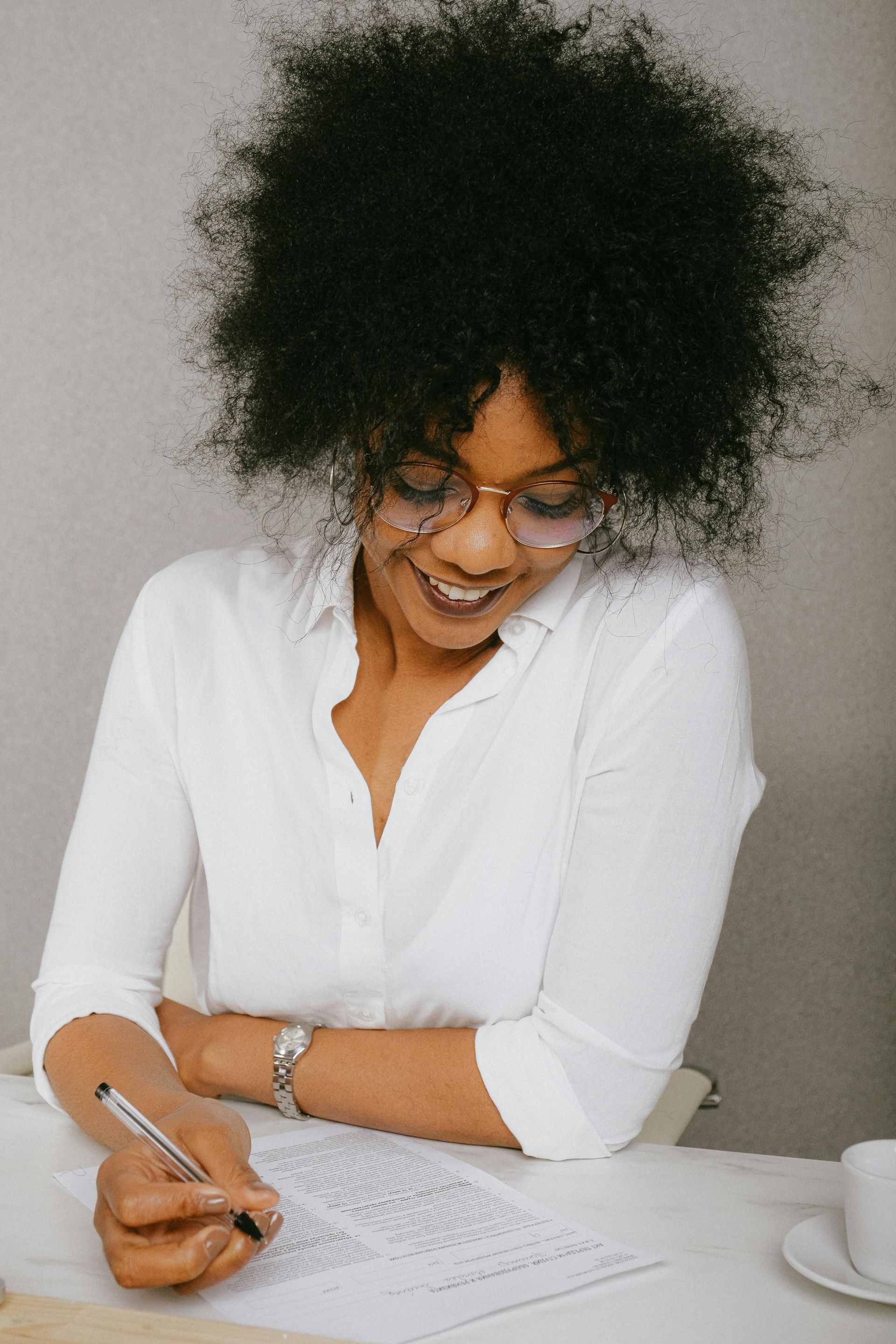 Woman with large afro smiling, writing on paper at a table. Wearing glasses and white shirt.
