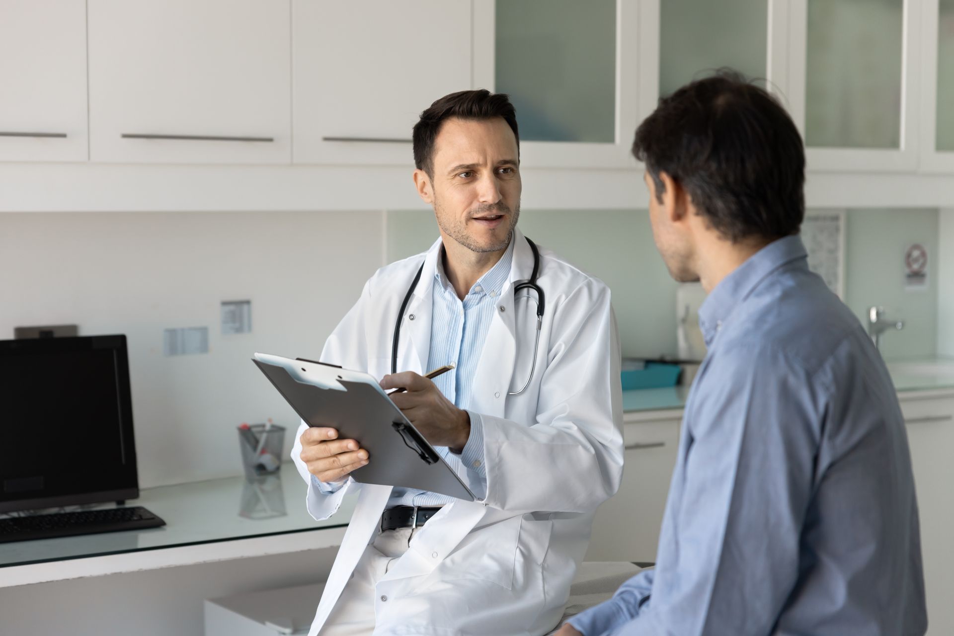 Doctor in white coat consults with a patient in a medical office, holding clipboard.