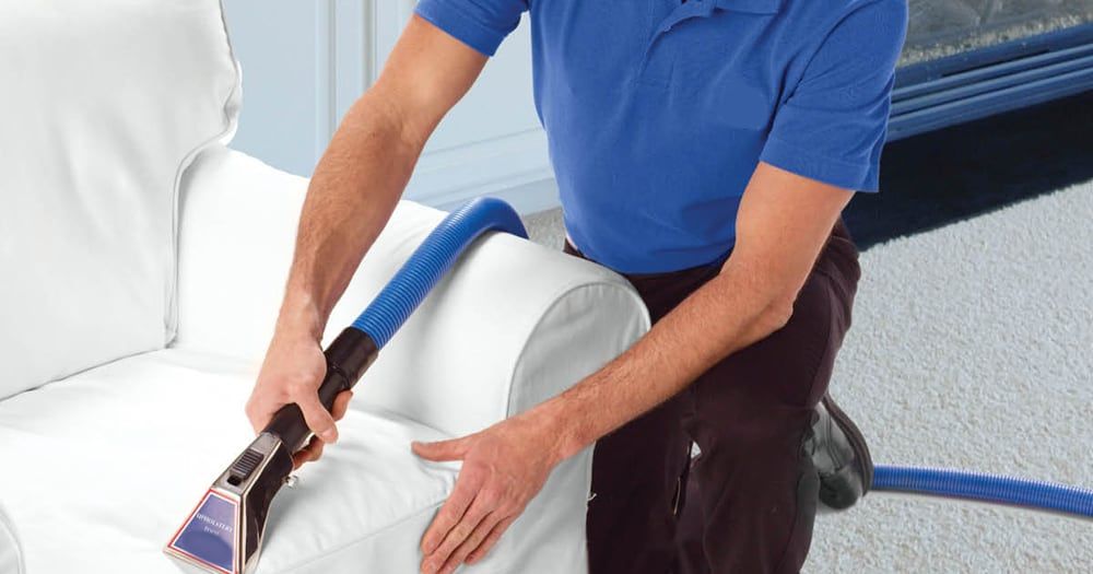 A person in a blue shirt uses an upholstery cleaning machine to clean a white armchair.