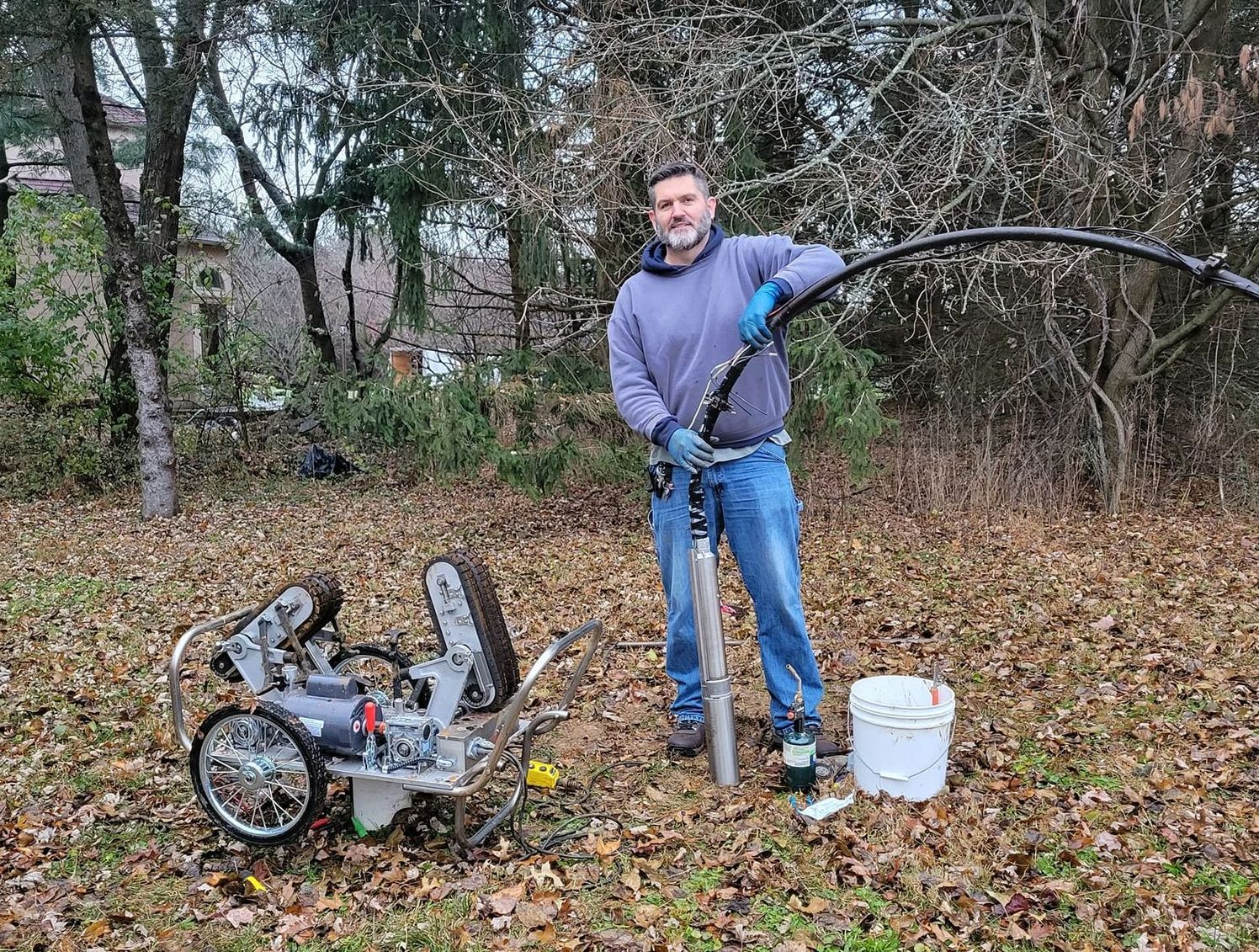 Man with equipment, likely drilling or pumping water, in a yard with leaves.