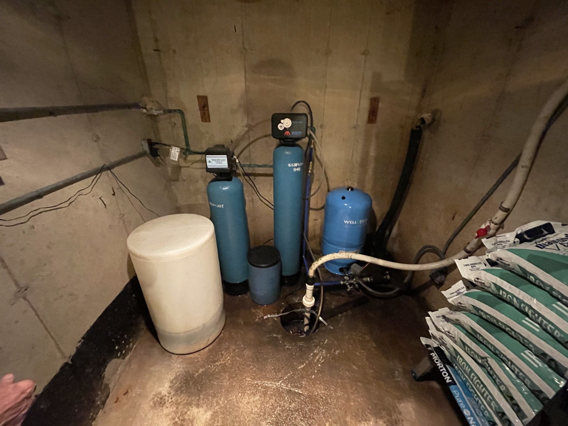 Water filtration system in a basement. Blue tanks, white salt container, and plumbing against concrete walls.