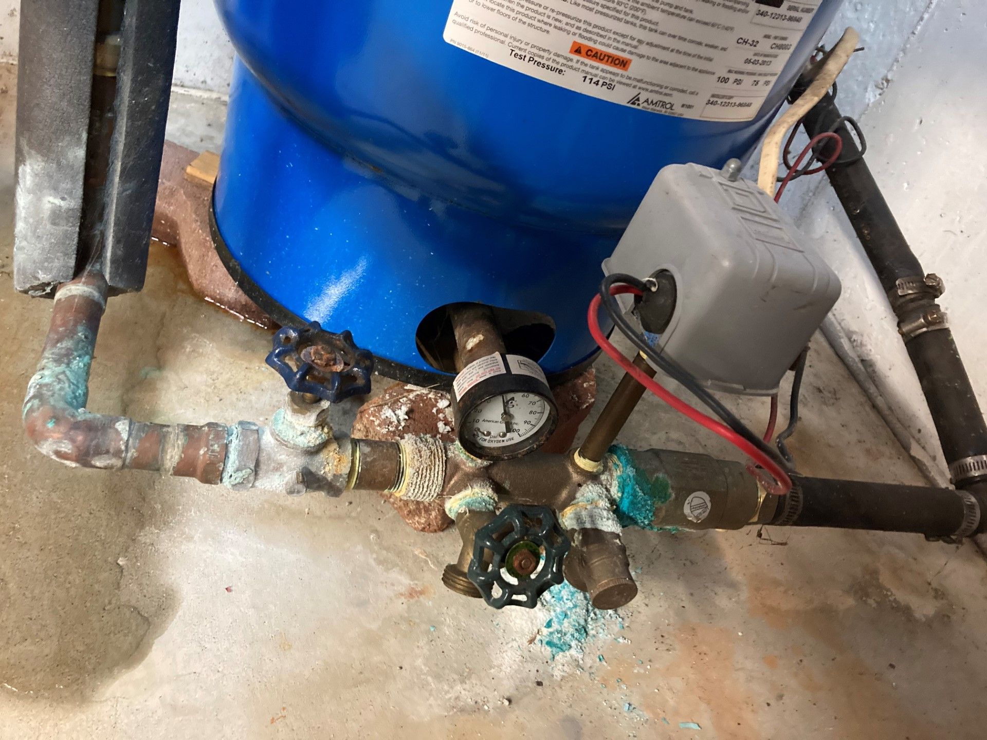 Blue water tank with pressure gauge and valves, showing signs of corrosion on the pipes.