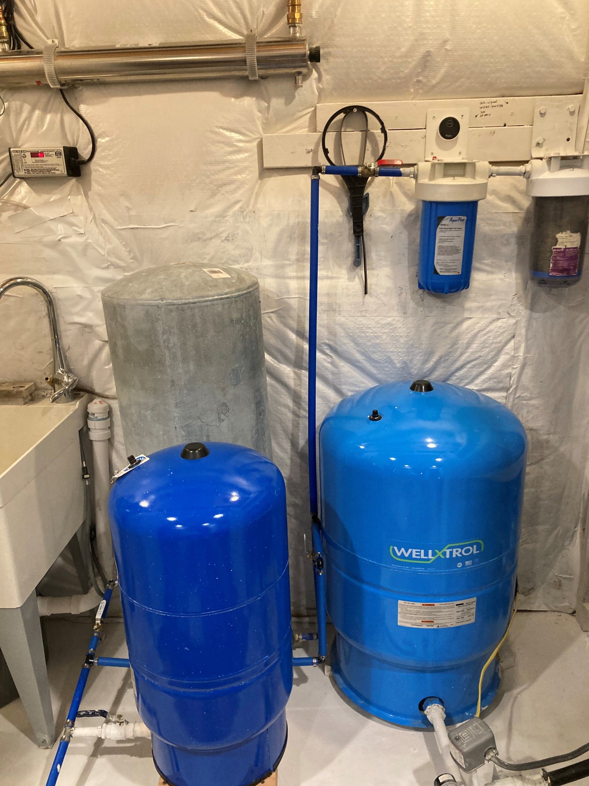 Water filtration and pressure tanks in a utility room. Blue and gray tanks, white walls, plumbing.