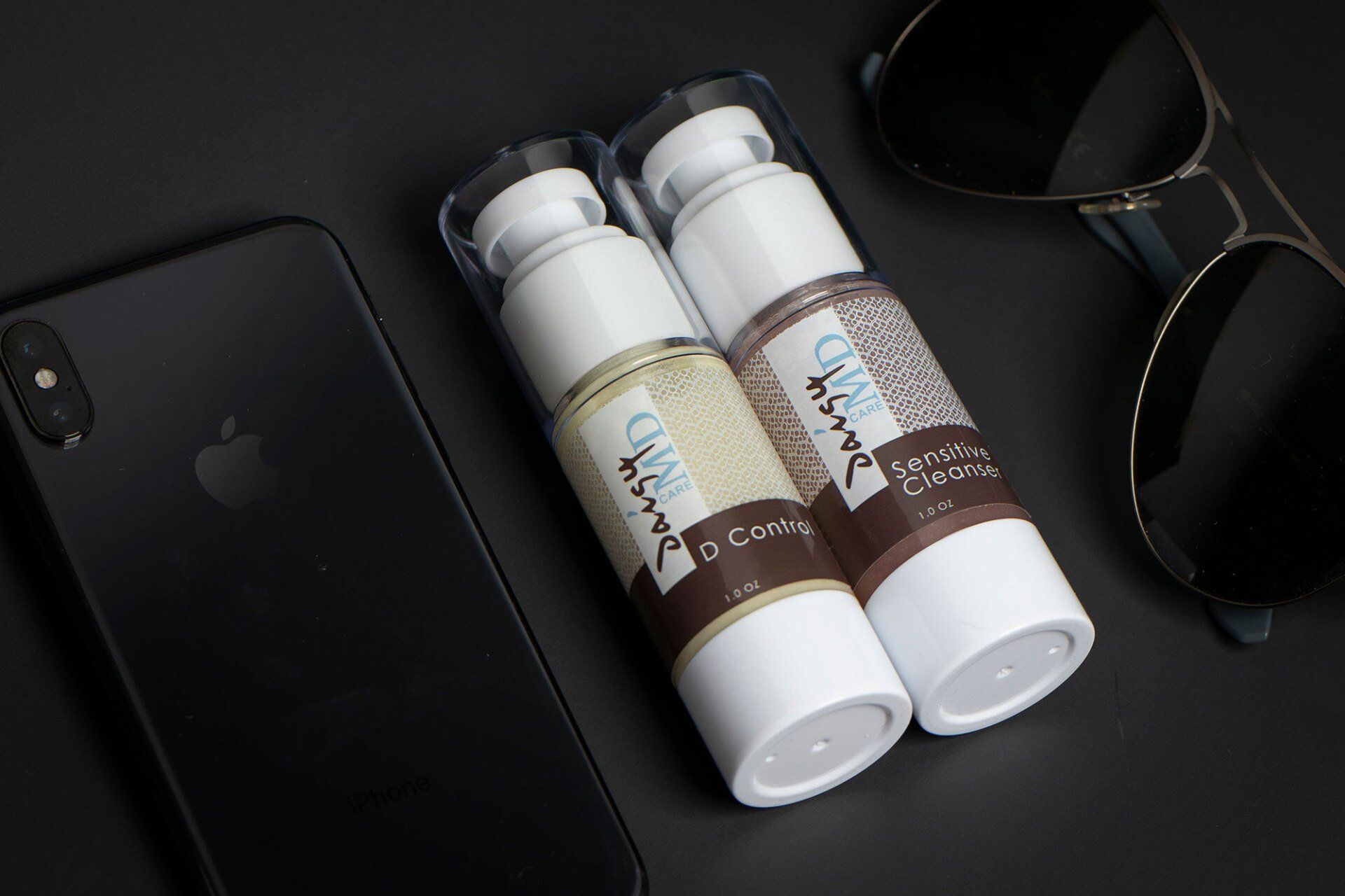 Two skincare bottles, phone, and sunglasses on a dark surface.