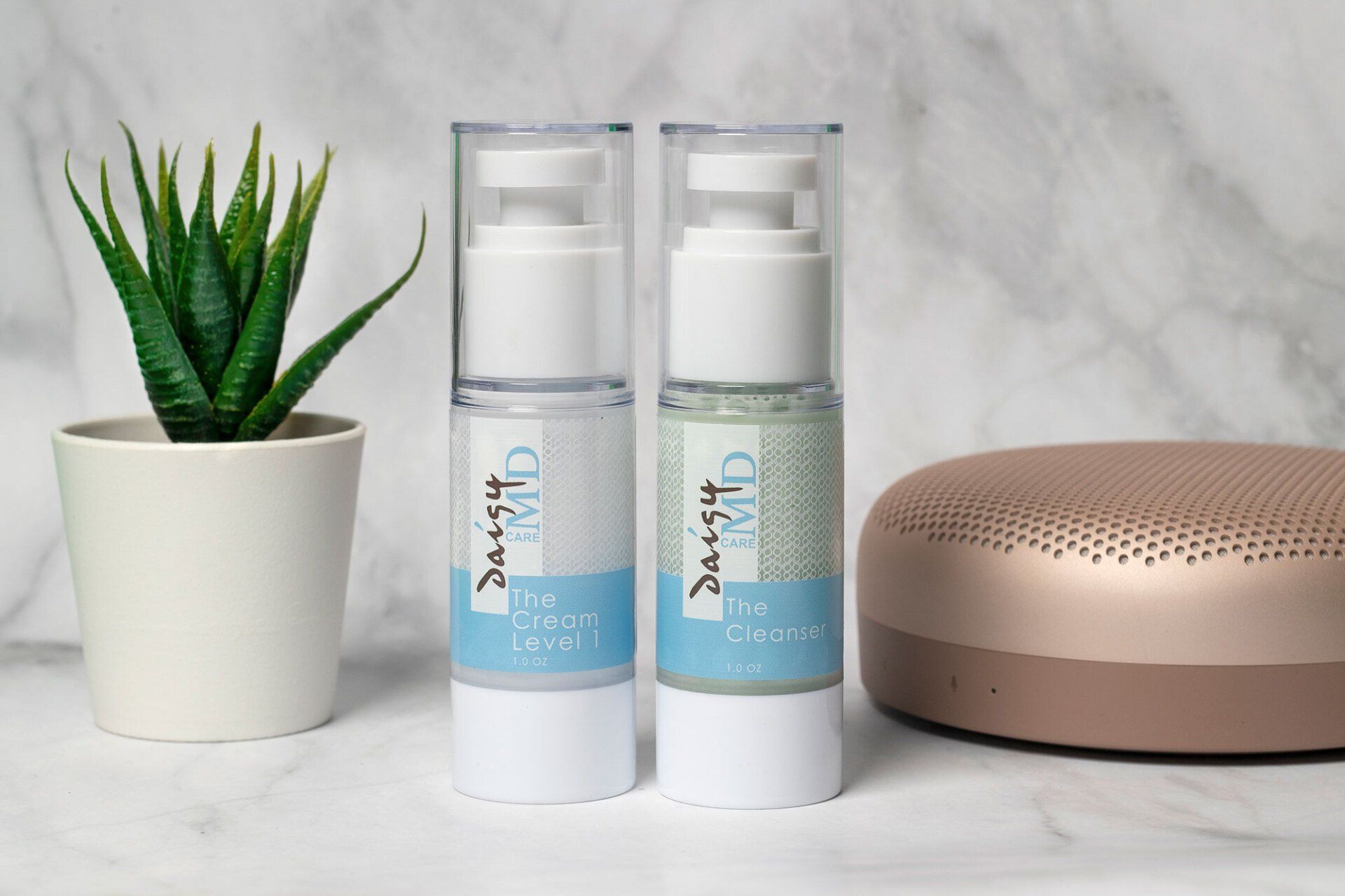 Two white cosmetic bottles, one green-tinted, beside a potted plant and speaker on a white surface.