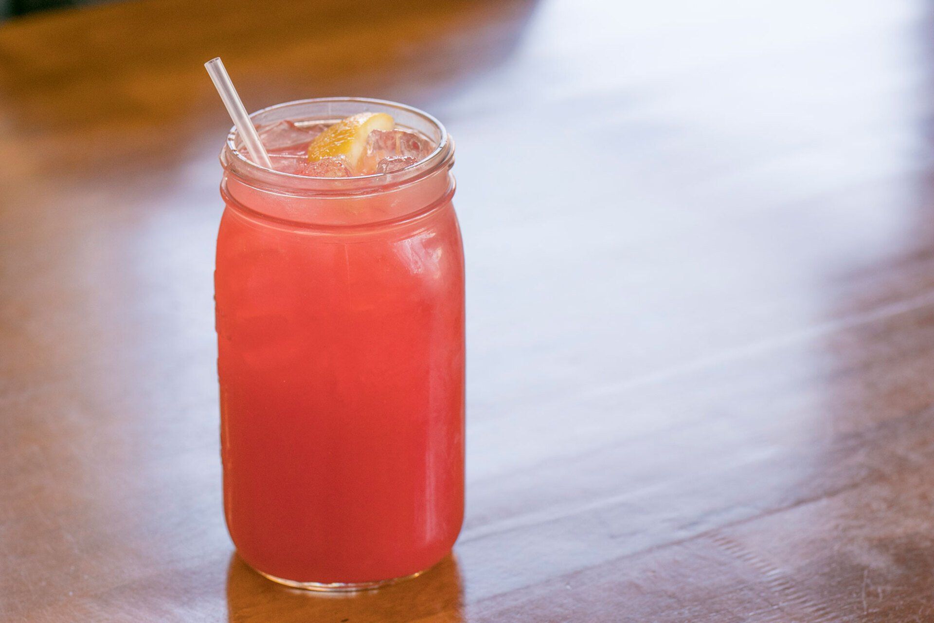 Pink beverage in a mason jar with a straw and lemon slice on a wooden table.