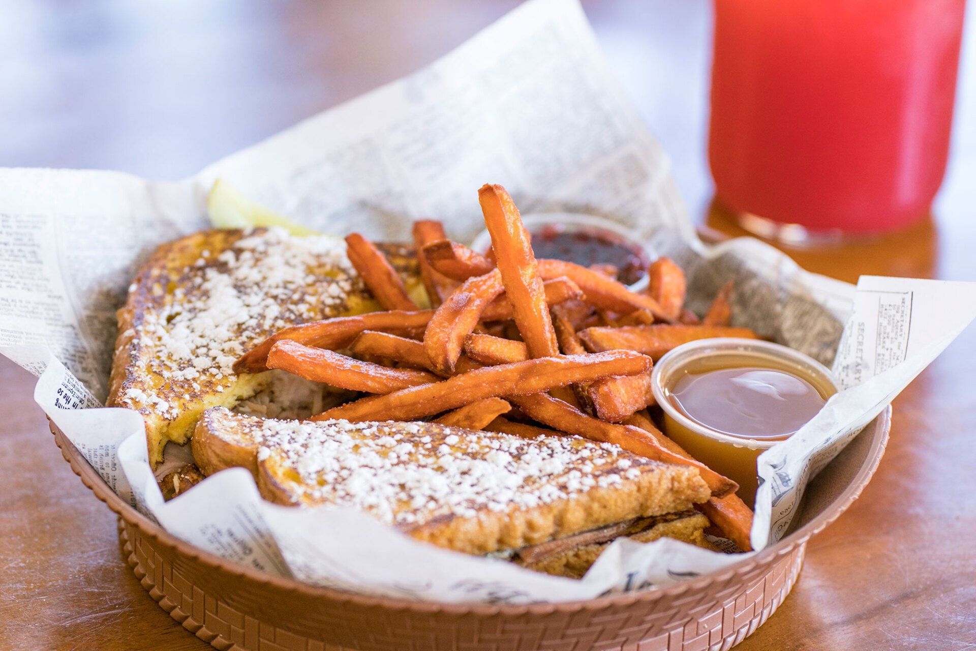 French toast, sweet potato fries, and sauces served in a basket, with a red drink in the background.
