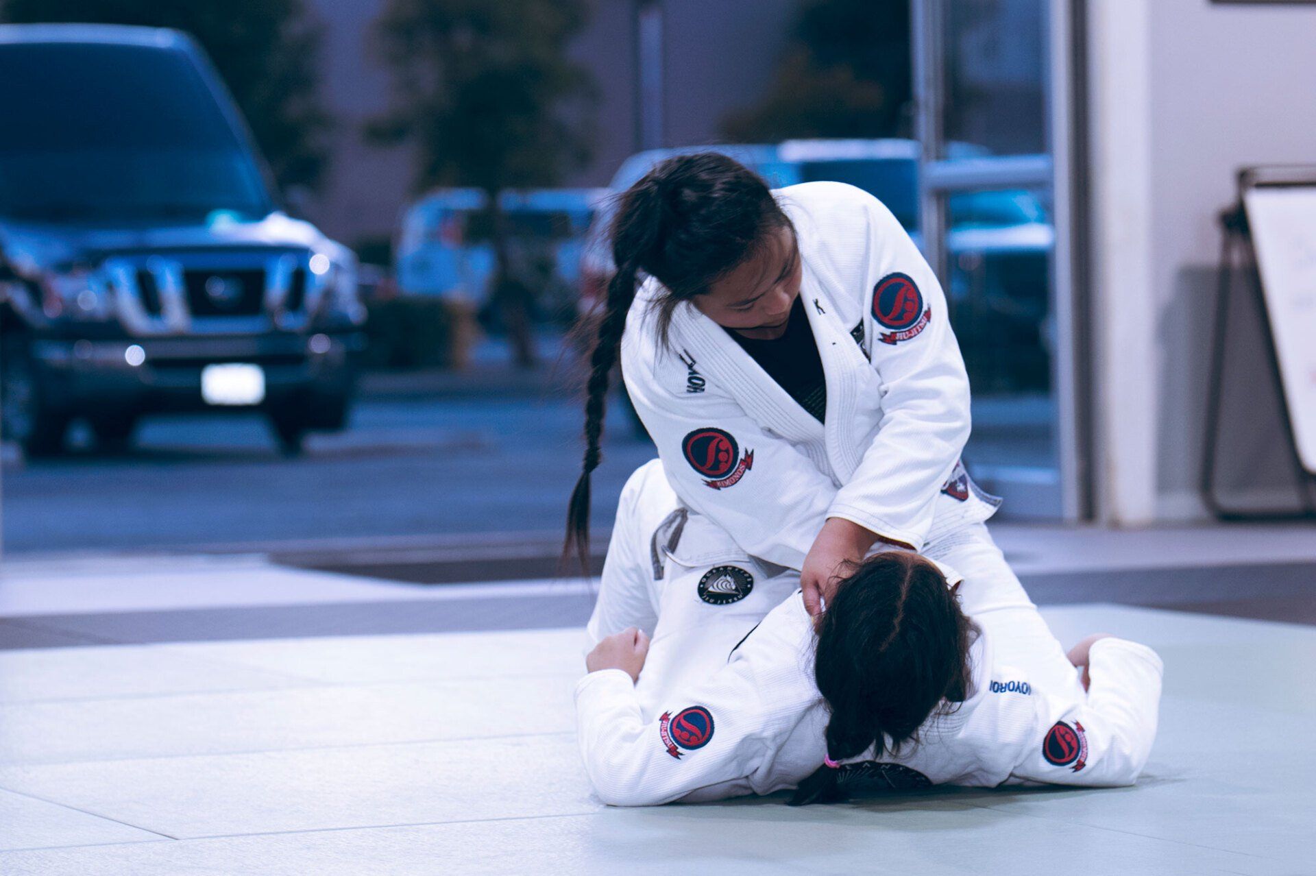 Two people in white martial arts uniforms practice a grappling move indoors.