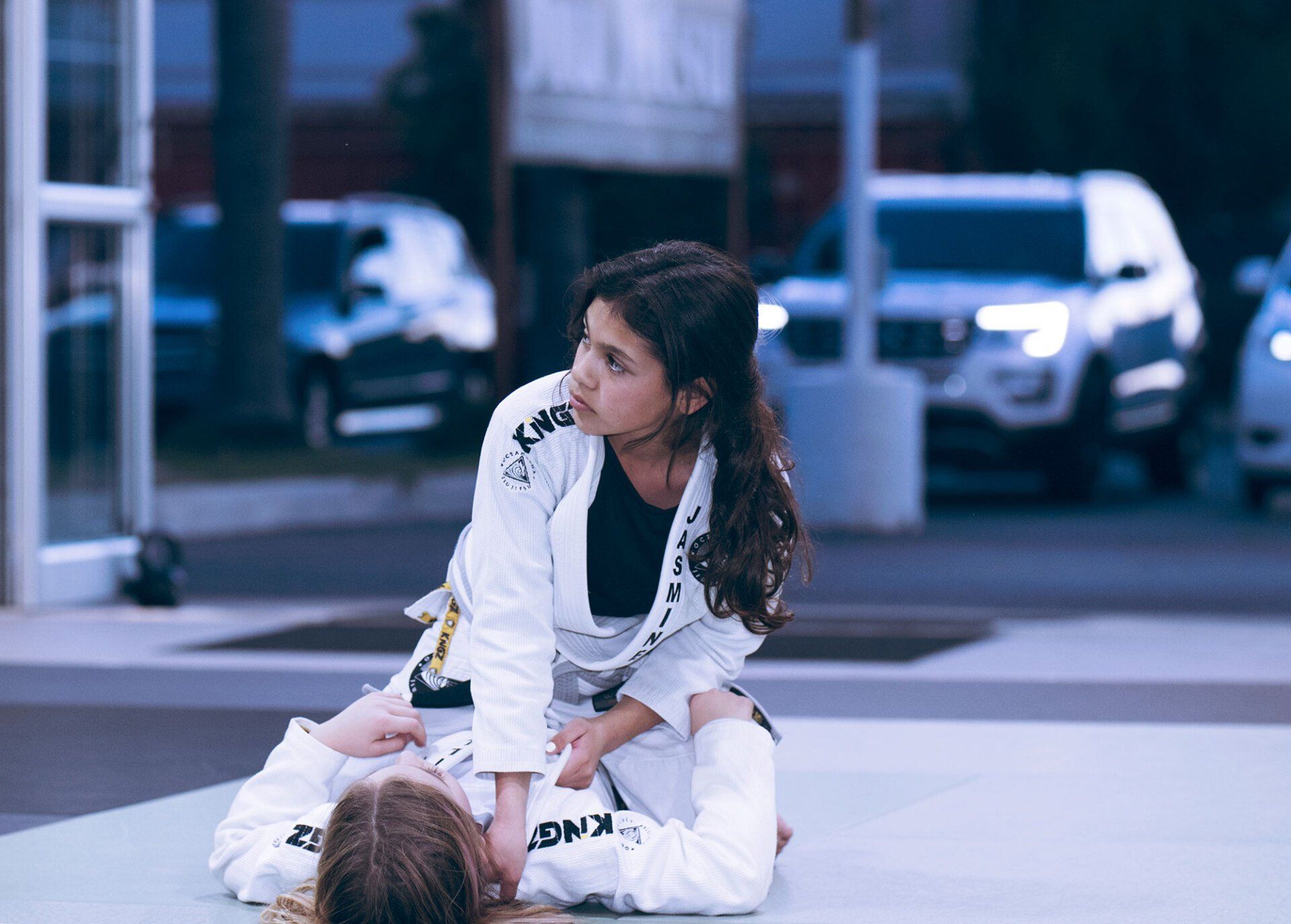 Two people in white martial arts uniforms practice on a mat, outdoors, with cars in the background.