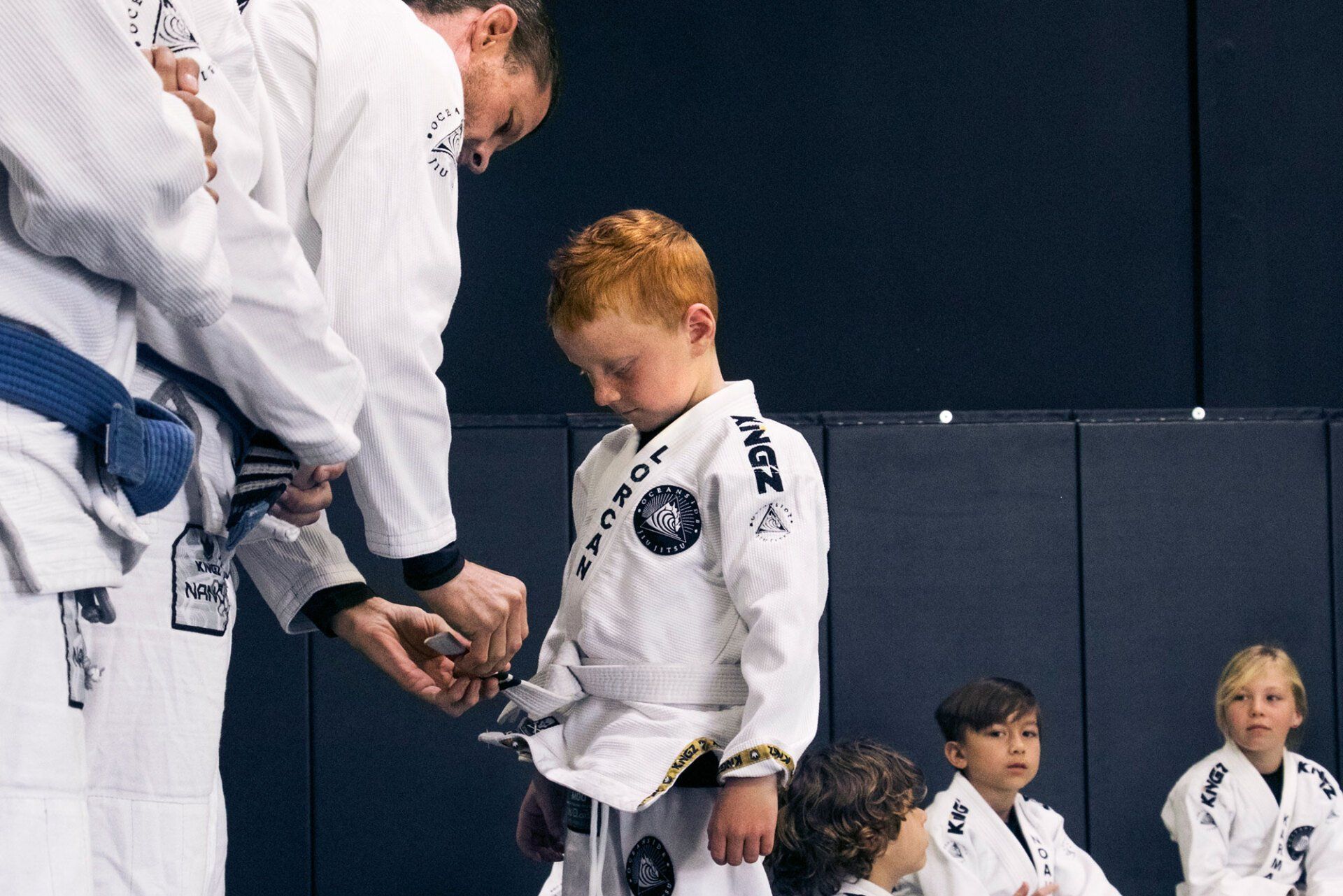 A boy in a white gi having his belt tied by an adult at a martial arts studio. Other children watch.