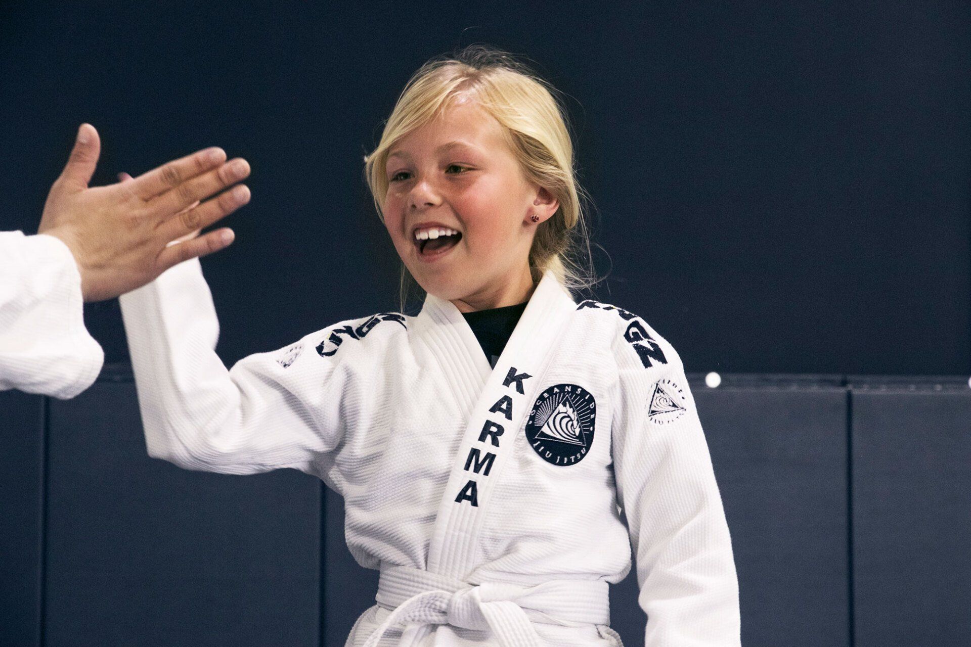 Young person in a white martial arts uniform, smiling, high-fiving someone out of frame.