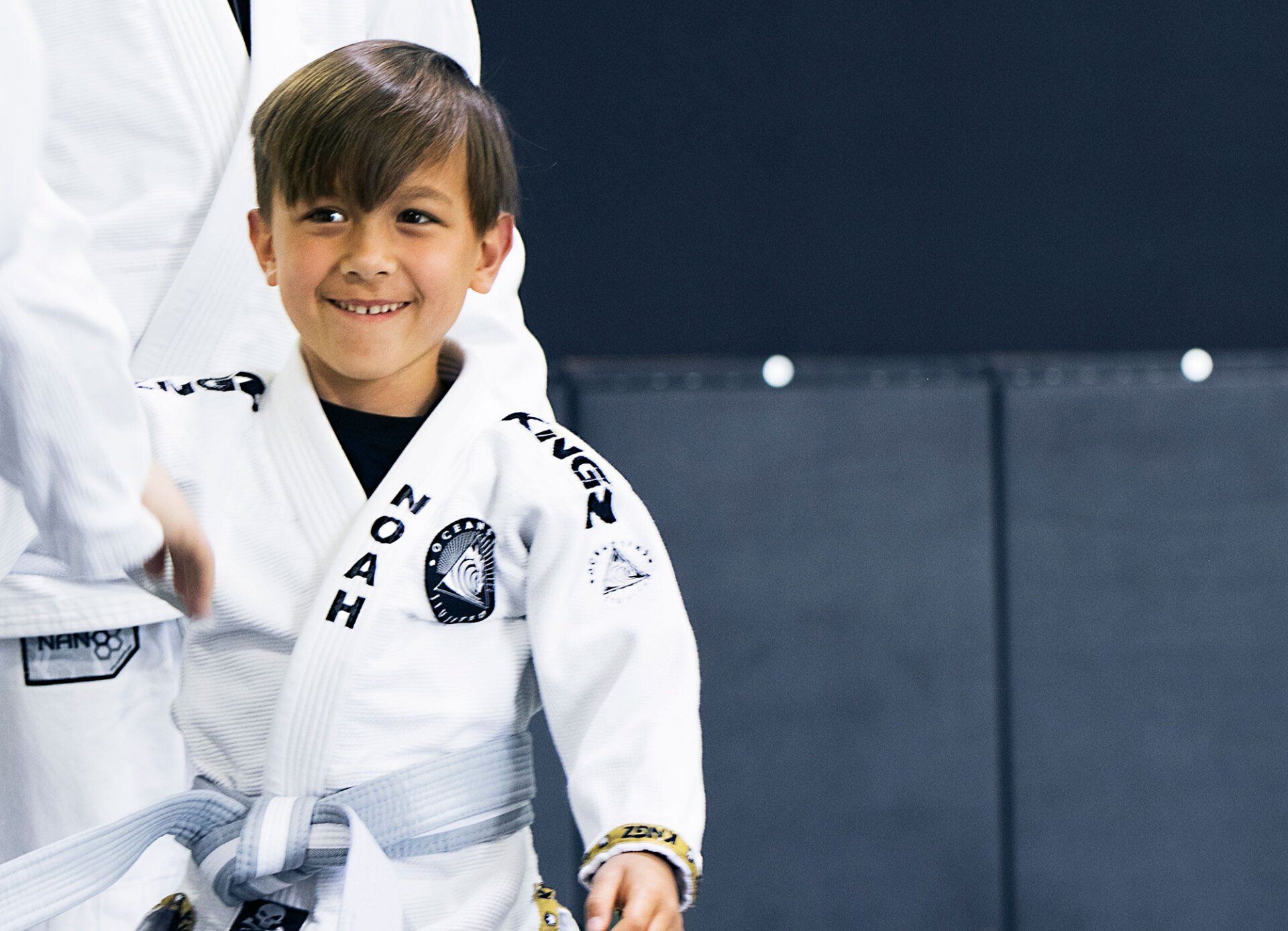 Smiling child in a white gi with a gray belt, standing in a martial arts setting.