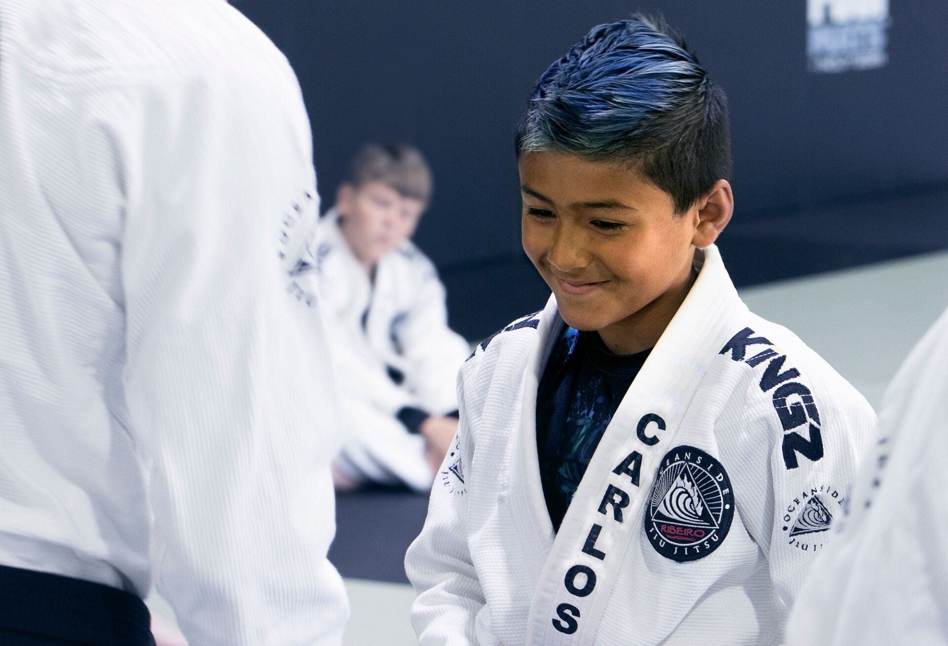 Boy in white martial arts uniform with blue-dyed hair smiles, in a training gym.